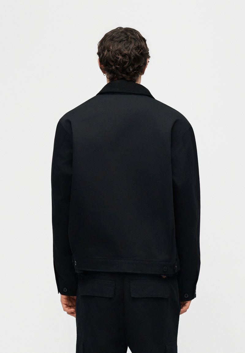 Mann mit dunklen lockigen Haaren, der eine schlichte schwarze Jacke und schwarze Hose trägt, von hinten vor weißem Hintergrund gezeigt.