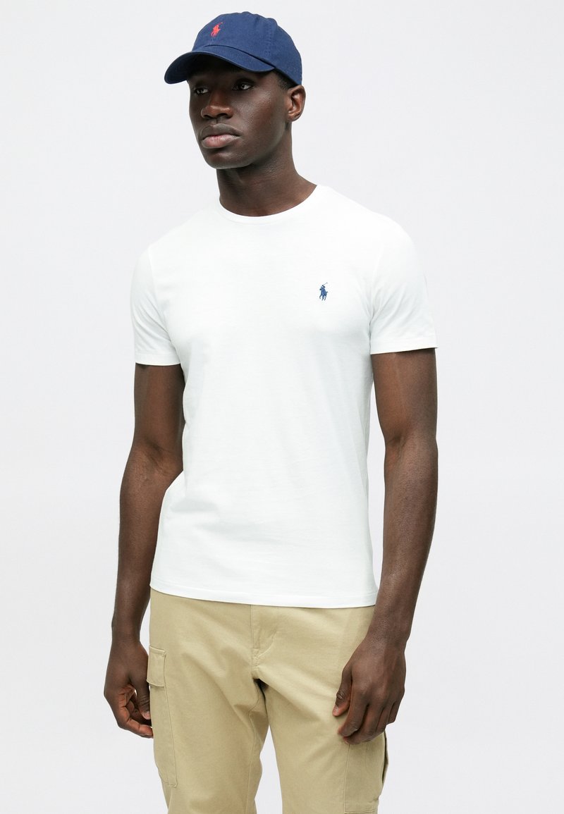 T-shirt bianca a maniche corte con un piccolo logo blu sul petto, abbinata a pantaloni cargo color cachi e un berretto blu navy. Design semplice e casual.