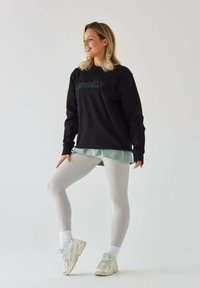 Czarna bluza z napisem "Księżniczka Pilates", nałożona na jasnoniebieską koszulę, w połączeniu z jasnoszarymi legginsami i białymi trampkami.