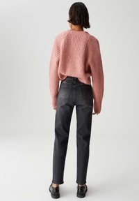 Donna che indossa un maglione rosa a larghe maglie, jeans neri a vita alta e scarpe nere, in piedi di spalle su uno sfondo chiaro.