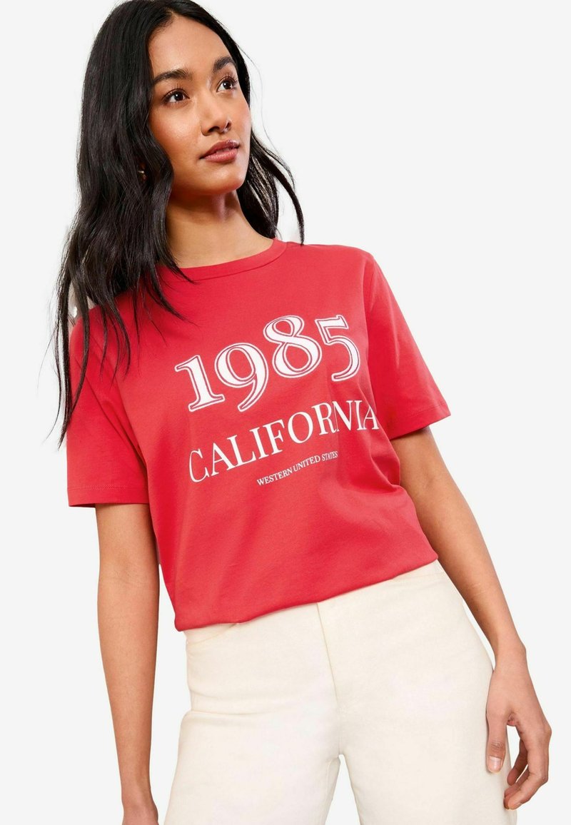 Punainen puuvillapaita, jossa on pyöreä kaula-aukko, lyhyet hihat ja valkoinen painatus, jossa lukee "1985 CALIFORNIA WESTERN UNITED STATES." Rennon mallinen.