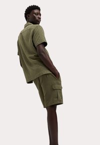 Camisa de manga corta color verde oliva con una textura sutil, combinada con pantalones cortos cargo a juego que cuentan con bolsillos laterales y un ajuste relajado.