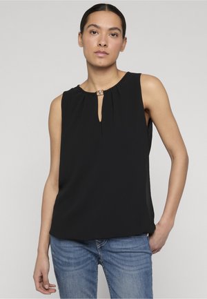 Femeie cu părul castaniu prins la spate, purtând un top negru fără mâneci cu decolteu tip cheie și blugi albaștri, stând în fața unui fundal simplu.