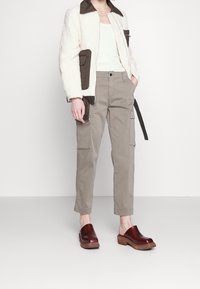 Bílá fleecová bunda s texturou a hnědými akcenty, šedé cargo kalhoty s bočními kapsami a kaštanové dřevěné klobouky.