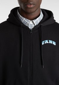 Czarna bluza z kapturem na zamku błyskawicznym z troczkami, z jasnoniebieskim logo "VANS". Nosi ją na wierzchu koszuli w kratę z kołnierzykiem w niebiesko-białym kolorze.