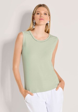 Vrouw draagt een mouwloze lichtgroene top en witte broek, met grote heldere edelsteenoorringen, poseert met handen in de zakken tegen een lichte achtergrond.