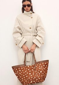 Smeđa torba s bijelim mrljama, s glatkim kožnim detaljima i dvostrukim naramenicama, prikazana uz svijetlu bež baloner.