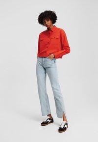 Rode cropped shirt met twee borstzakken, gecombineerd met lichtblauwe recht gesneden jeans met zijsplitten, en zwarte sneakers met witte accenten.