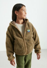 Giacca a zip in pile marrone con cappuccio, caratterizzata da un rivestimento beige, superficie testurizzata e una toppa con dettagli multicolori. Indossata sopra un top chiaro.