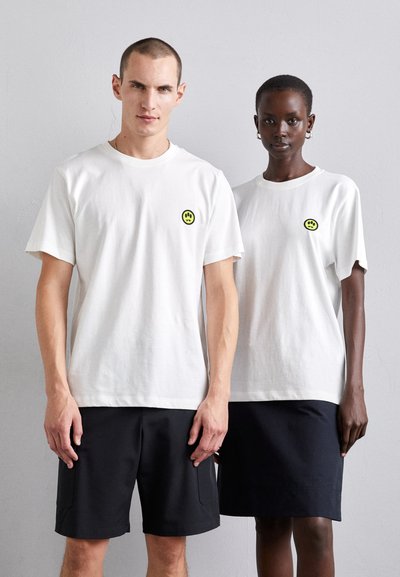 Des t-shirts en coton blancs avec des cols ronds, arborant de petits logos de smiley jaunes sur la poitrine gauche, associés à des shorts et des jupes noirs.