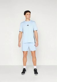 Αθλητική μπλε γαλάζια μπλούζα και σορτς με μαύρες λεπτομέρειες και λογότυπο. Το μοντέλο φοράει μαύρα αθλητικά παπούτσια και λευκές κάλτσες, στέκεται όρθιο.