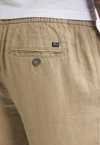 Pantalon en lin beige avec un tissu texturé. Doté d'une poche arrière avec un bouton et une étiquette logo. Ceinture  élastique pour un confort optimal.