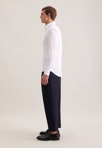 Biała koszula z długim rękawem i kołnierzykiem, w parze z ciemnogranatowymi, dopasowanymi spodniami i czarnymi butami, widziana z boku na neutralnym tle.