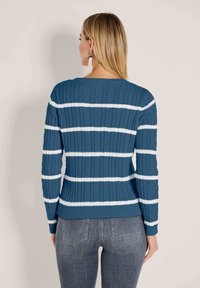 Frau, die von hinten zu sehen ist, trägt einen blauen Pulli mit Zopfmuster und weißen horizontalen Streifen sowie graue Jeans und steht vor einem schlichten beigen Hintergrund.