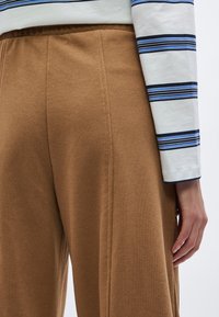 Bruine, zachte stoffen broek met een elastische tailleband, getoond naast een wit shirt met blauwe en marineblauwe strepen.