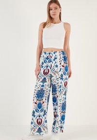 Losse, wijde broek van witte stof met een levendig blauw en rood bloemenpatroon. Gecombineerd met een aansluitend wit crop top en witte sneakers.