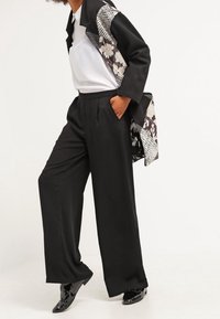 Pantalons larges noirs associés à un haut blanc. La veste noire présente des motifs floraux et pied-de-poule, avec des chaussures noires brillantes.