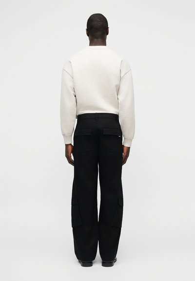 Άνδρας με λευκό πουκάμισο μακρυμάνικο και μαύρο χαλαρό παντελόνι cargo με τσέπες με καπάκια, στέκεται με την πλάτη προς το μέρος μας πάνω σε απλό φωτεινό φόντο.