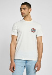 Koszulka z krótkim rękawem, kremowa, wykonana z bawełny, z niebieskim i czerwonym logo Lee Jeans na lewej piersi. Prosty, klasyczny design z okrągłym dekoltem.