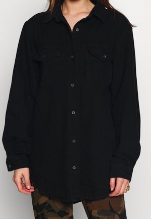 Kobieta ubrana w luźną czarną koszulę na guziki z kieszeniami na piersi oraz w spodnie moro, stojąca na prostym jasnym tle.