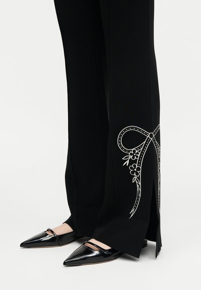 Κάτω μέρη ποδιών με μαύρο παντελόνι με λευκό κέντημα φλοράλ φιόγκων και πλαϊνές σχισμές, σε συνδυασμό με γυαλιστερά μαύρα παπούτσια Mary Jane με μυτερή μύτη.