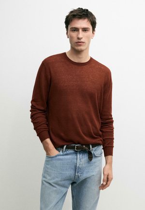 Giovane uomo che indossa un maglione a maniche lunghe color ruggine, jeans azzurri chiari e una cintura intrecciata nera, in piedi contro uno sfondo bianco semplice.