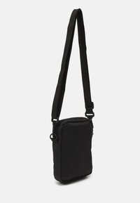 Sac bandoulière noir en tissu doux, de forme rectangulaire, avec une sangle réglable et un dessus zippé. Aucun motif ni logo visible.
