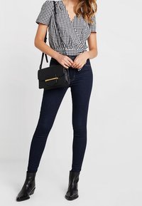 Top con diseño de cuadros en blanco y negro, con mangas cortas, combinado con jeans ajustados azul oscuro y botas de tobillo negras. Pequeño bolso cruzado negro.