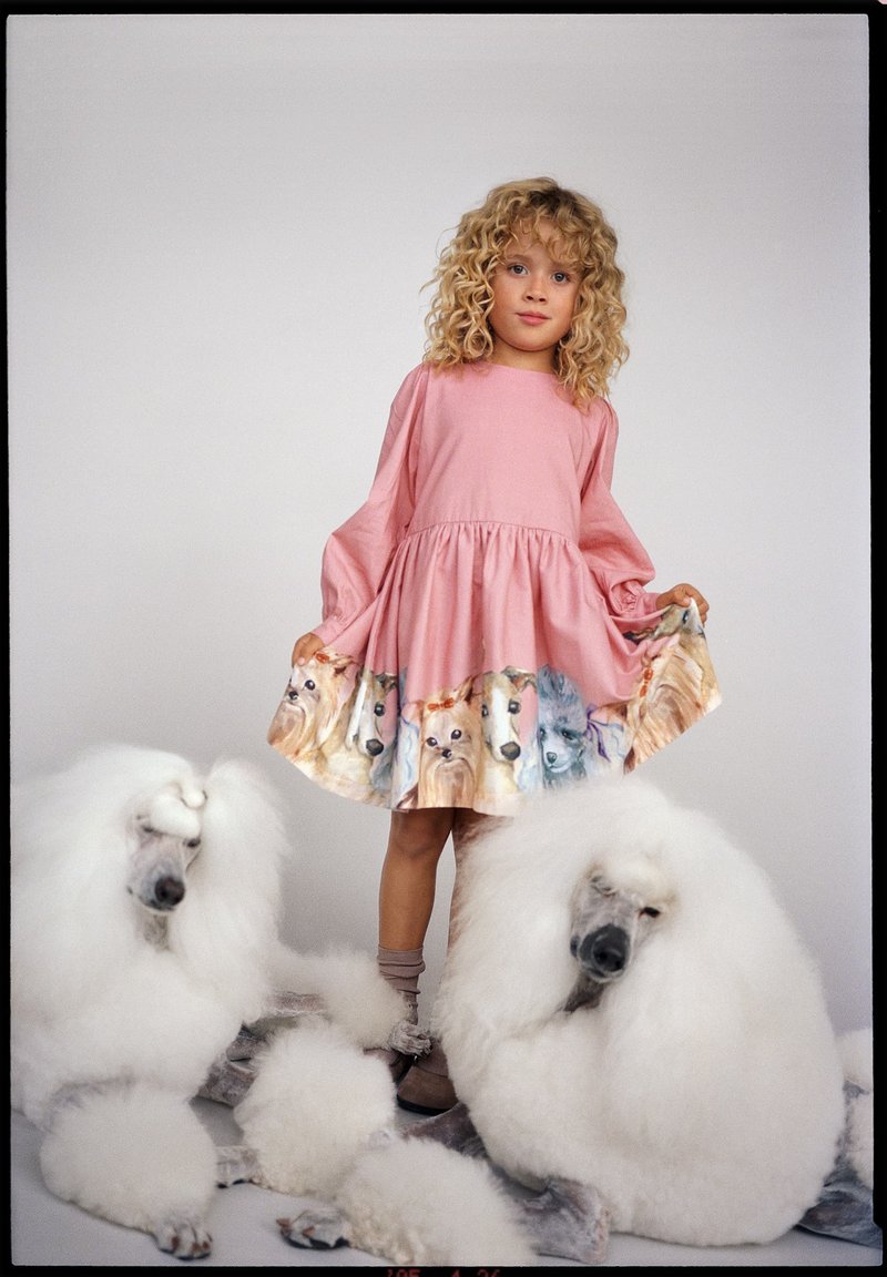 Jeune fille en robe rose avec des illustrations de chiens se tient entre deux grands caniches blancs sur un fond uni.