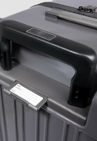 Grau Hartschalenkoffer mit strukturierter Oberfläche, schwarzem Kunststoffgriff, Gepäckanhängerhalter und Reißverschluss oben.