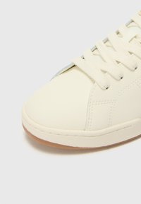 White leather sneaker featuring a rounded toe, flat cream laces, and a gum rubber sole. Subtle stitching and grey accents near the laces.