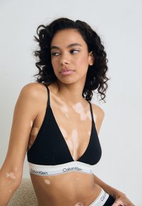 Bralette triangulaire noir avec des bretelles fines, featuring une bande élastique blanche avec le logo "Calvin Klein". Tissu lisse, design minimaliste.