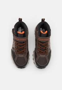 Zapatillas deportivas marrones con acentos negros, parte superior de malla texturizada, cordones naranjas, cuello acolchado y una lengüeta trasera para facilitarlas al ponérselas.