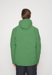 Persona che indossa una giacca con cappuccio verde e pantaloni scuri, voltata di spalle contro uno sfondo chiaro e uniforme.