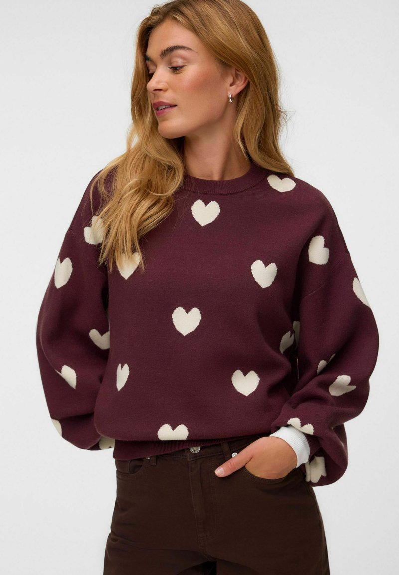 Femme aux longs cheveux blonds portant un pull bordeaux avec des motifs de cœurs blancs et un pantalon foncé, posant avec une main dans la poche.