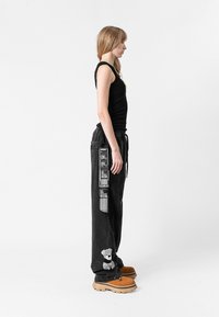 Pantalones cargo de mezclilla negra con parches laterales, cintura con cordón y bordado de oso; combinados con zapatos marrones de cordones; modelo posando de lado.