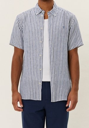 Mand iført en kortærmet, blå og hvid vertikalt striped skjorte over en hvid T-shirt med mørkeblå bukser.