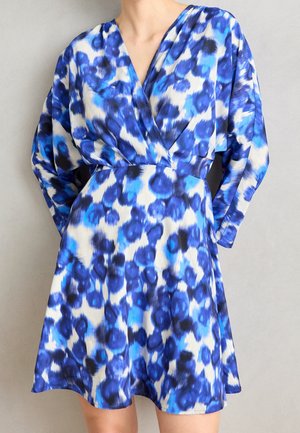 Frau trägt ein blau-weißes, langärmeliges Wickelkleid mit Blumenmuster, V-Ausschnitt und tailliertem Bund, steht vor einem schlichten Hintergrund.