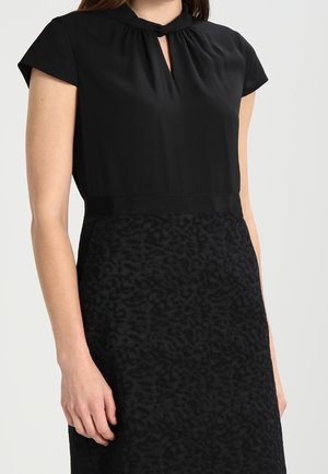 Kvinde iført en sort kjole med kappelignende ærmer, en vridd nøglehul-udskæring og en subtil mønstret nederdelssektion.