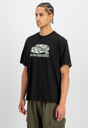 Giovane uomo con capelli intrecciati che indossa una t-shirt nera Alpha Industries con una grafica di un veicolo militare e pantaloni cargo verde oliva.