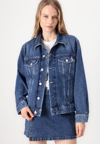 Denim jacket in dark blue, featuring a classic collar, button front, two chest pockets, and distressed texture with visible stitching details.