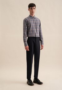 Camicia a scacchi con bottoni blu, bianca e rossa con maniche lunghe, abbinata a pantaloni navy aderenti e scarpe in pelle nere. Sfondo chiaro.