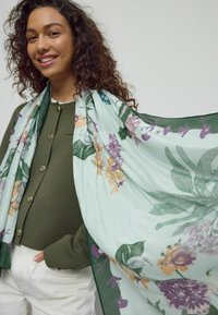 Kwiatowy szal w jasnym miętowym odcieniu z purpurowymi, pomarańczowymi i zielonymi wzorami. Lekka tkanina opada na modelkę ubrana w oliwkową zieloną bluzkę.