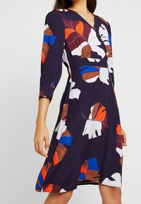 Marineblaues Kleid mit V-Ausschnitt und Dreiviertelärmeln, das ein lebhaftes Blattmuster in Rot, Blau, Orange, Braun und Weiß aufweist, mit lockerer Passform.