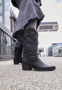 Černé kovbojské boty s texturovaným designem a středním podpatkem, spárované se roztrhanou šedou sukní. Povrchy jsou hladké s matným povrchem.