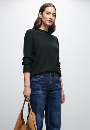 Maglione a costine verde scuro con maniche rimboccate, abbinato a jeans di denim blu; modella che tiene una borsa a mano in pelle beige.