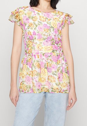 Kobieta ubrana w białą bluzkę z krótkim rękawem w żółty, różowy i fioletowy wzór kwiatowy, zestawioną z jasnoniebieskimi dżinsami.