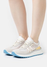Baskets de sport légères en daim beige et en maille, avec des accents jaunes, une semelle en caoutchouc bleue et un système de laçage.