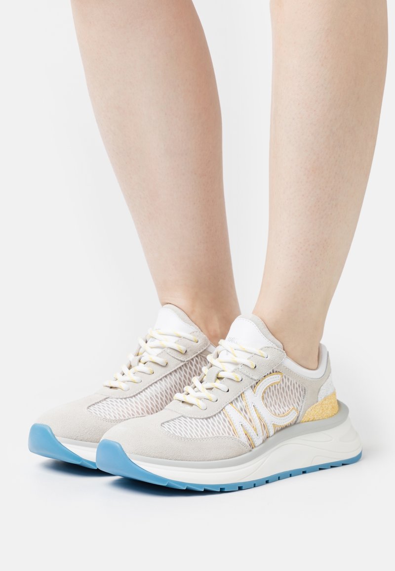 Baskets de sport légères en daim beige et en maille, avec des accents jaunes, une semelle en caoutchouc bleue et un système de laçage.