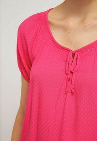 Blouse rose à manches courtes en tissu texturé, col rond, détail noué et bouton décoratif à l'avant. Conception douce et légère.